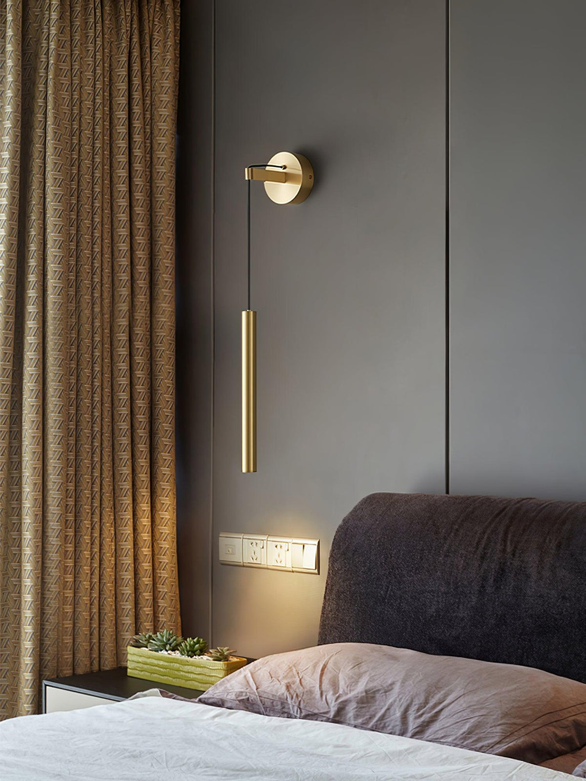 Brass wall light hangs in a bedroom with a brown textured curtain and a grey headboard, a warm glow beneath, from Clérachic.