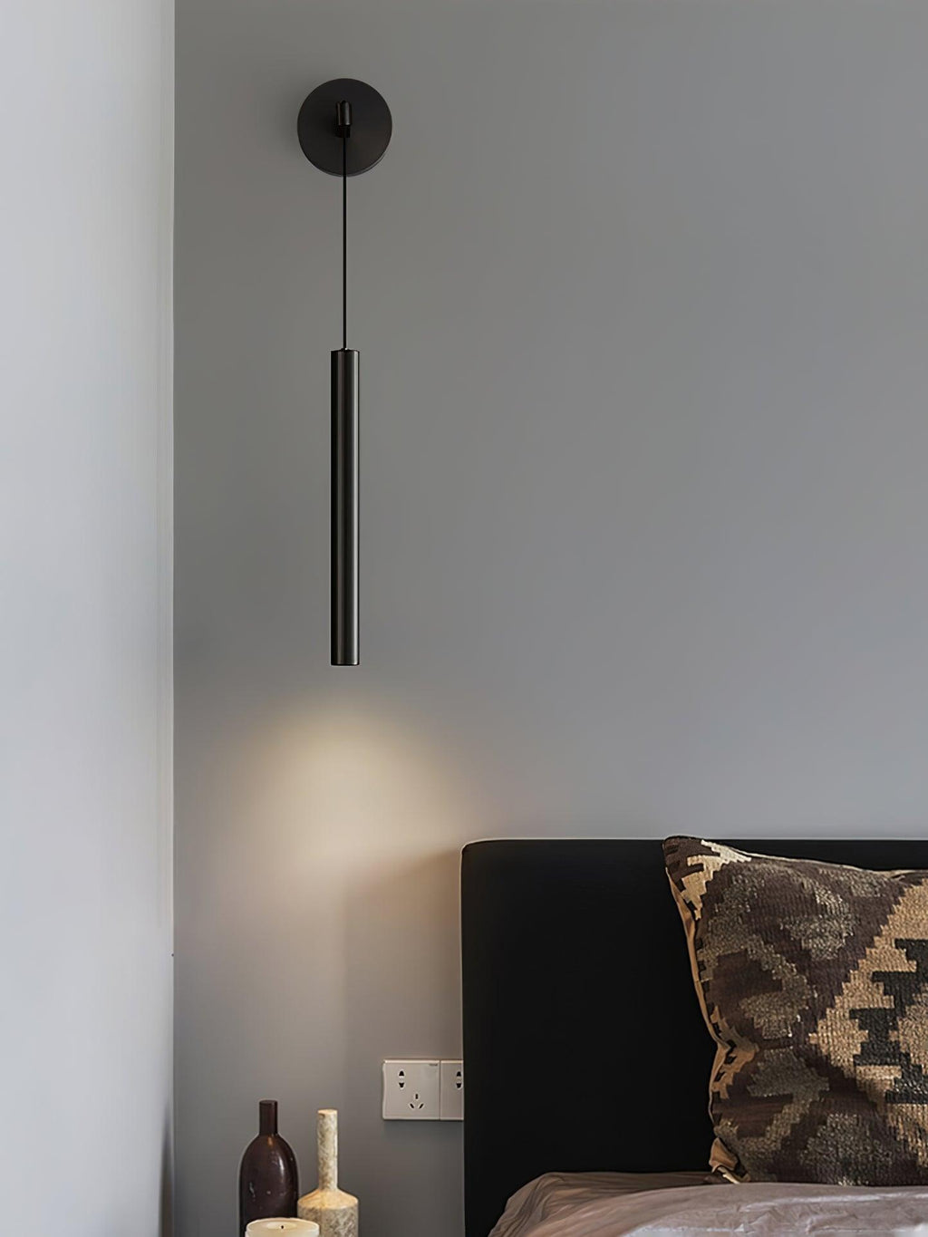 Black wall light above a bed with a woven patterned cushion and glass bottles on the side table, a Clérachic wall light.