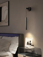Black tube wall light above a bedside table with a framed photo, next to a framed print and a dark headboard, from Clérachic.