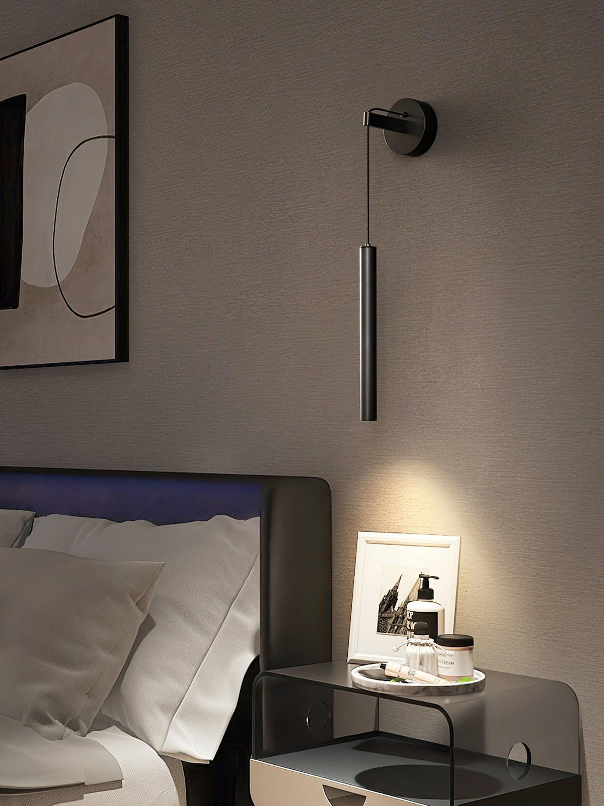 Black tube wall light above a bedside table with a framed photo, next to a framed print and a dark headboard, from Clérachic.