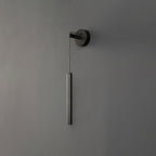 Black tube wall light on a dark grey wall, with its round backplate and cord, lit softly, from Clérachic.