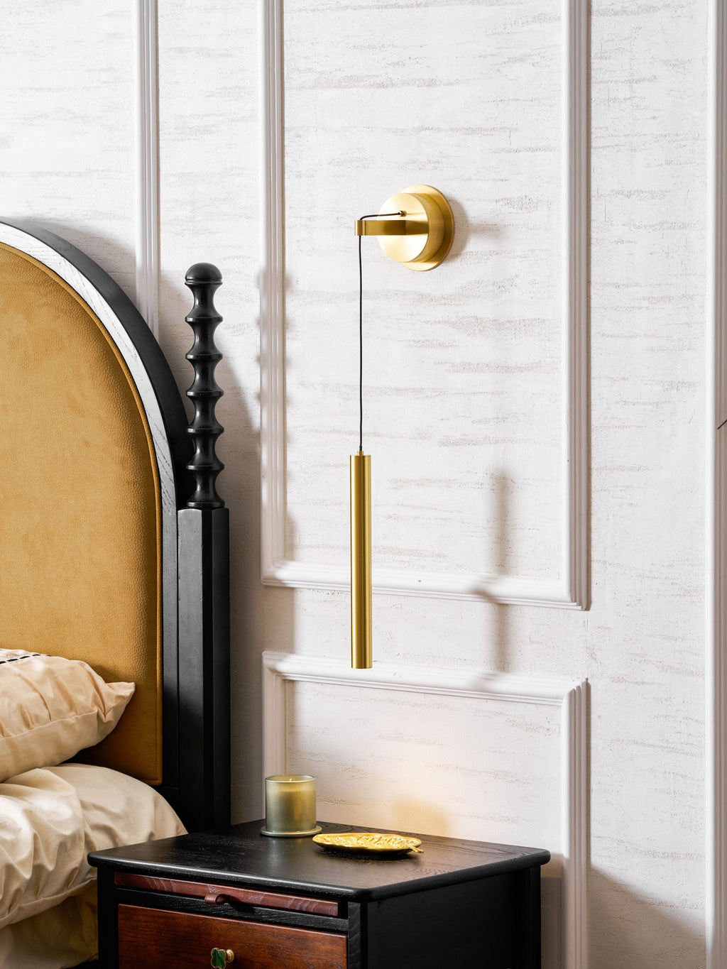 Brass wall light on a white panelled wall above a black bedside table with a candle and a gold dish, from Clérachic.