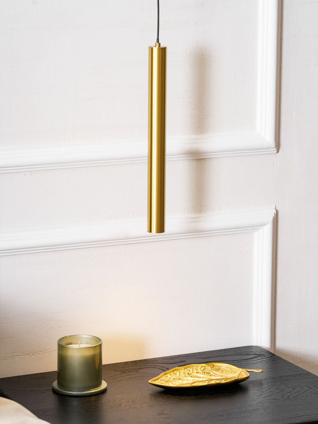 Brass tube hangs over a dark table with a green glass candle and a gold leaf dish, against white panelling, a Clérachic wall light.