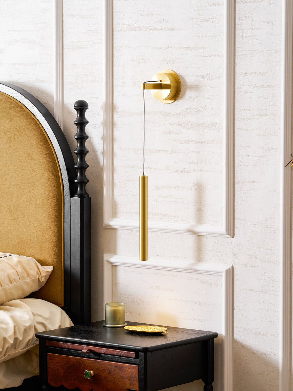 Brass wall light high on a panelled wall, with a velvet headboard and a black bedside table with a drawer, a Clérachic wall light.