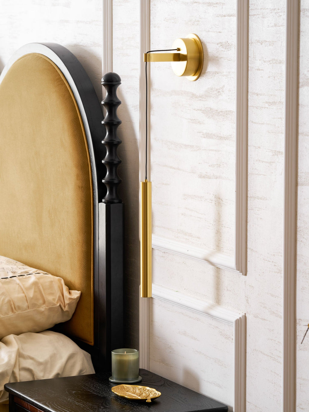 Side view of a brass wall light beside a velvet arched headboard, with a candle and gold dish on the table below, from Clérachic.