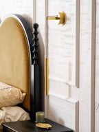 Side view of a brass wall light beside a velvet arched headboard, with a candle and gold dish on the table below, from Clérachic.