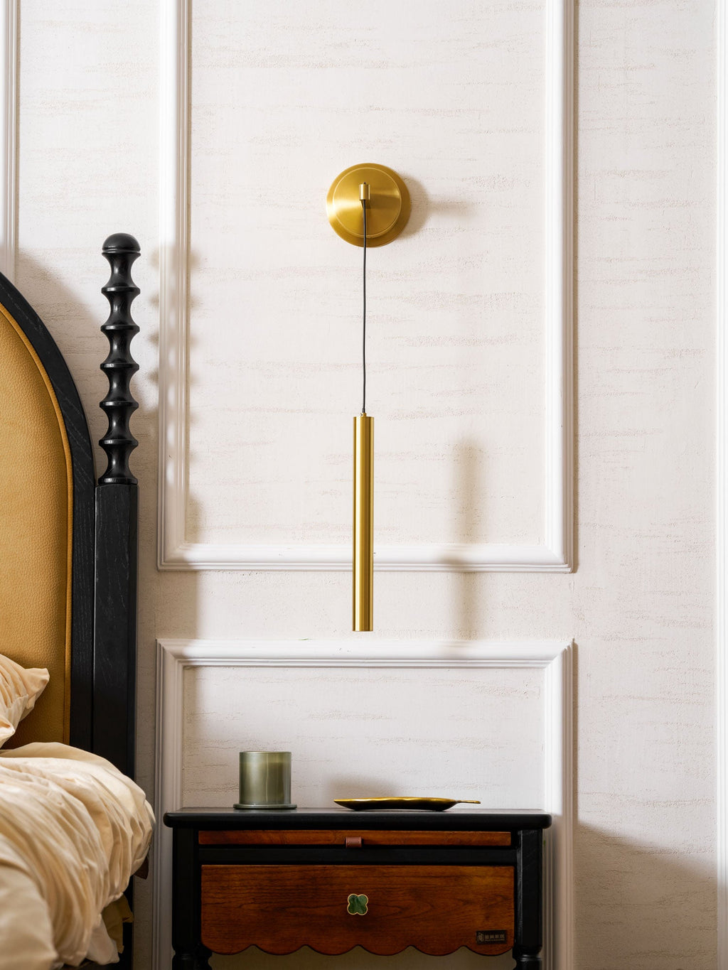 Brass wall light hanging over a bedside table with a drawer and a green candle, in a room with white wall panels, from Clérachic.