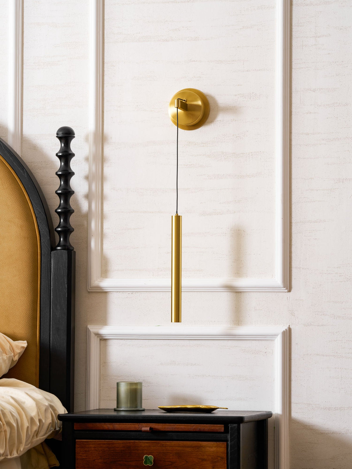 Brass wall light on a panelled wall next to a carved black bedpost, a candle glowing on the table below, a Clérachic wall light.