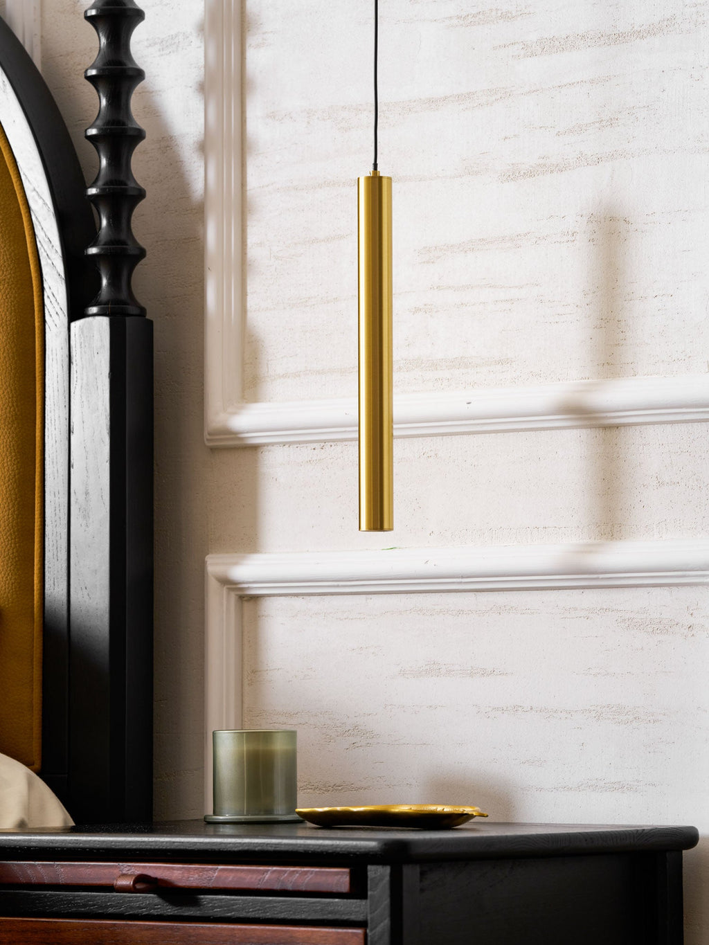 Lower section of a brass tube above a green glass candle and gold dish on a black bedside table, a Clérachic wall light.