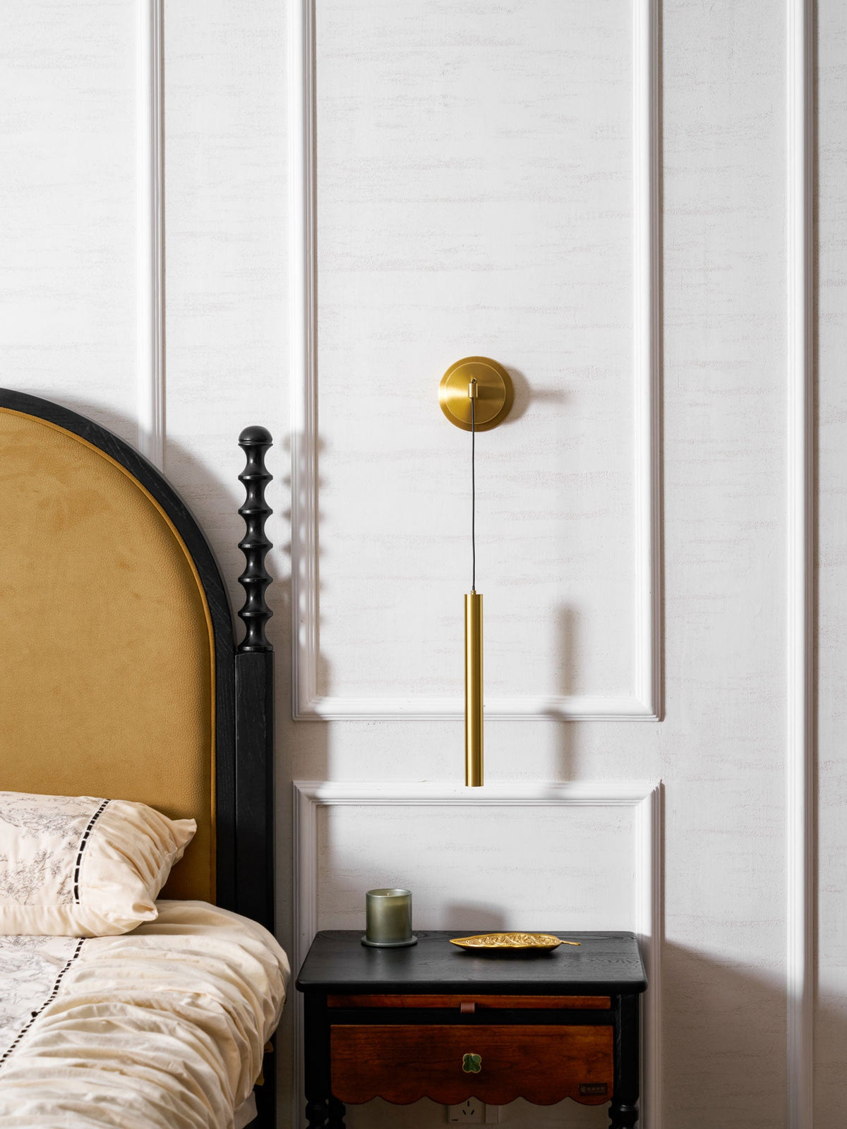 Brass wall light on a white panelled wall beside an arched headboard and a bedside table with a scalloped drawer, from Clérachic.