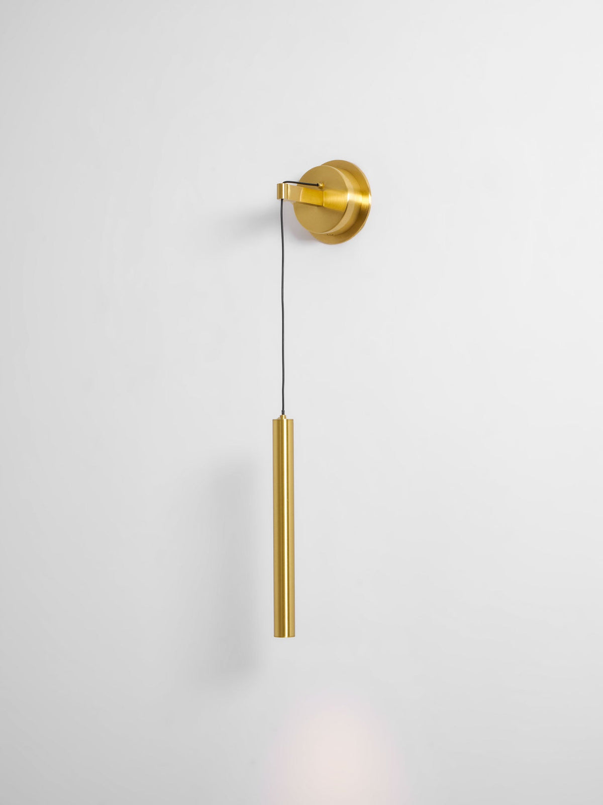 Brass wall light on a pale grey wall, with a faint glow of light below the tube, from Clérachic.