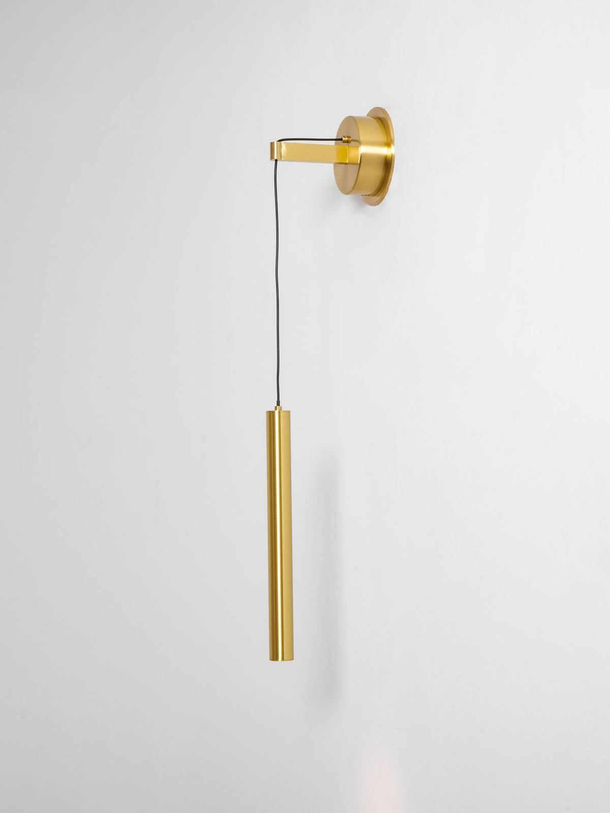 Brass wall light seen from a slight angle on a pale wall, showing the arm, cord and tube, a Clérachic wall light.