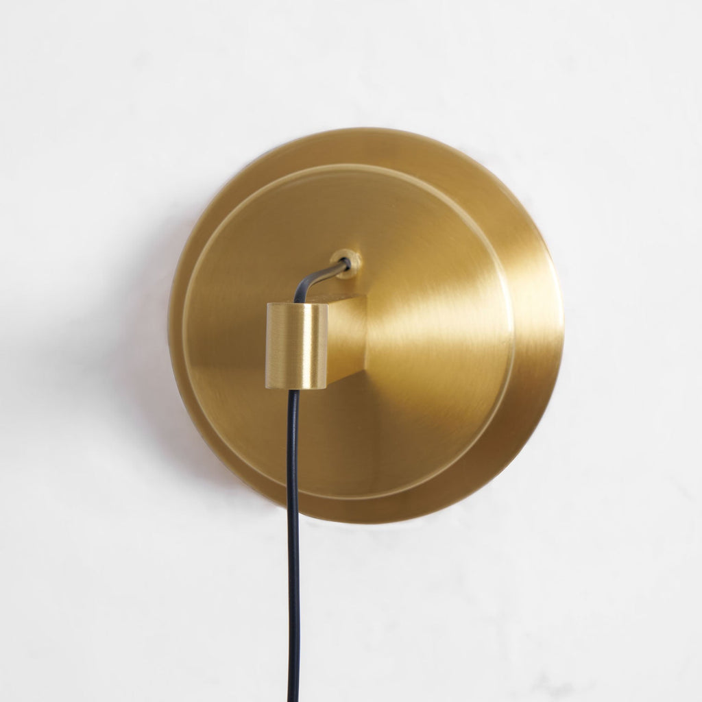 Close view of the round brass backplate with its stepped edge and a black cord leaving the arm, on this Clérachic wall light.