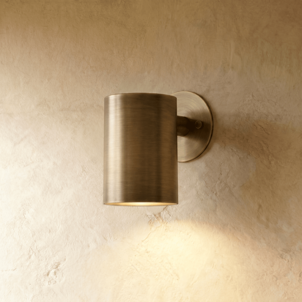 This Clérachic cylindrical wall light in aged vintage brass casts a warm downlight against a textured plaster wall.