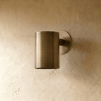A Clérachic cylinder wall light in aged vintage brass sits flush on a plaster wall, glowing softly from beneath.