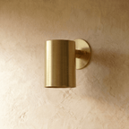 A Clérachic cylinder wall light in polished brass mounts flush to a plaster wall with light spilling downward.