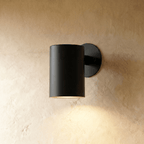 This Clérachic cylindrical wall light in matte black sits close to a plaster wall, downlight glowing beneath it.