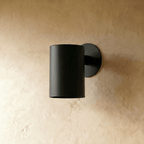 A Clérachic cylinder wall light in matte black mounts flush against a warm plaster surface, lit from below.
