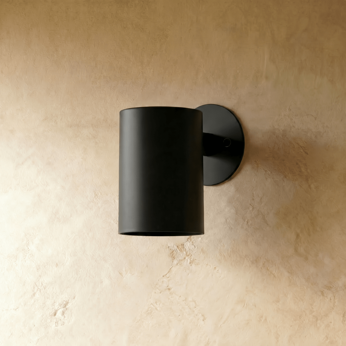 A Clérachic cylinder wall light in matte black mounts flush against a warm plaster surface, lit from below.
