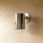 A Clérachic cylinder wall light in mirror-polished chrome sits flush on plaster, its downlight glowing softly.
