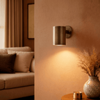 This Clérachic cylindrical wall light in aged vintage brass lights a living room corner beside a vase of dried grass.