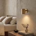 A Clérachic cylinder wall light in polished brass glows above a bedside table with books and a sprig of olive.
