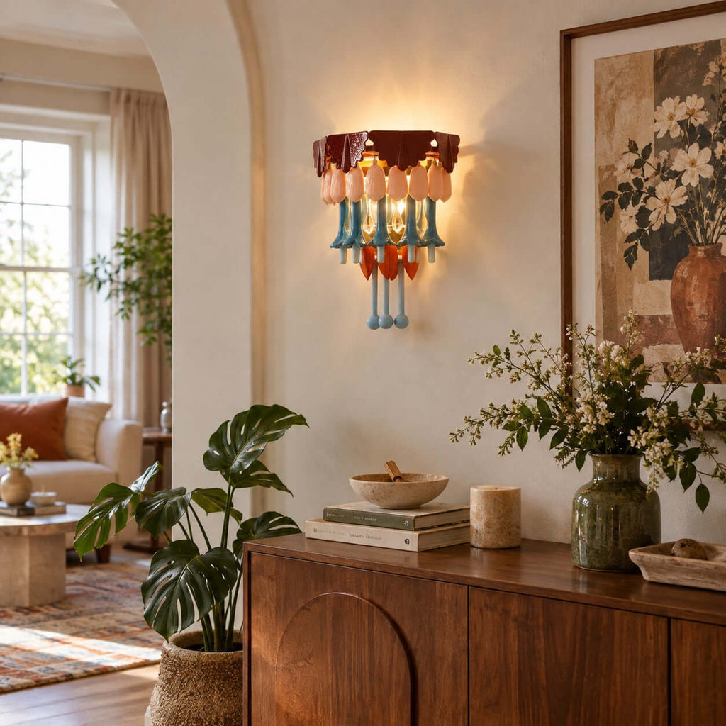 Clérachic wall sconce glowing above a walnut sideboard with a potted monstera, framed floral art and an archway to a sunlit room.