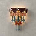 A Clérachic wall sconce lit against a grey plaster wall, showing its burgundy top, pink petals, teal stems and orange accents.