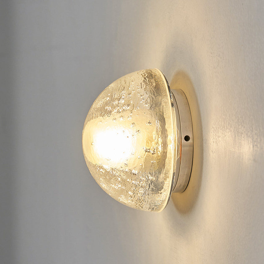 Side view of this Clérachic wall lamp glowing warm, its dome of clear bubbled glass casting a soft halo on a grey wall.