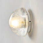 This Clérachic wall lamp seen unlit from the side, the clear dome of rippled, bubble-flecked glass sitting on a slim round plate.
