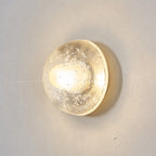 Front-on view of a Clérachic wall light switched on, its clear glass dome flecked with bubbles and glowing warm on a grey wall.