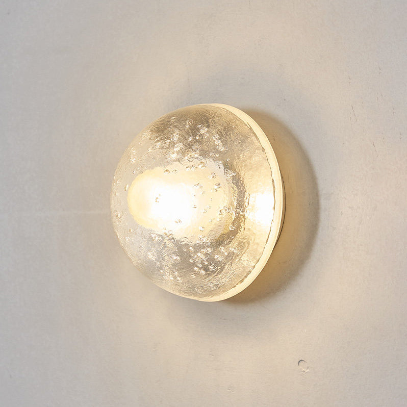 Front-on view of a Clérachic wall light switched on, its clear glass dome flecked with bubbles and glowing warm on a grey wall.