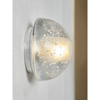 Angled view of a Clérachic wall lamp glowing warm beside a wooden panel, the clear dome sitting flush on its round plate.