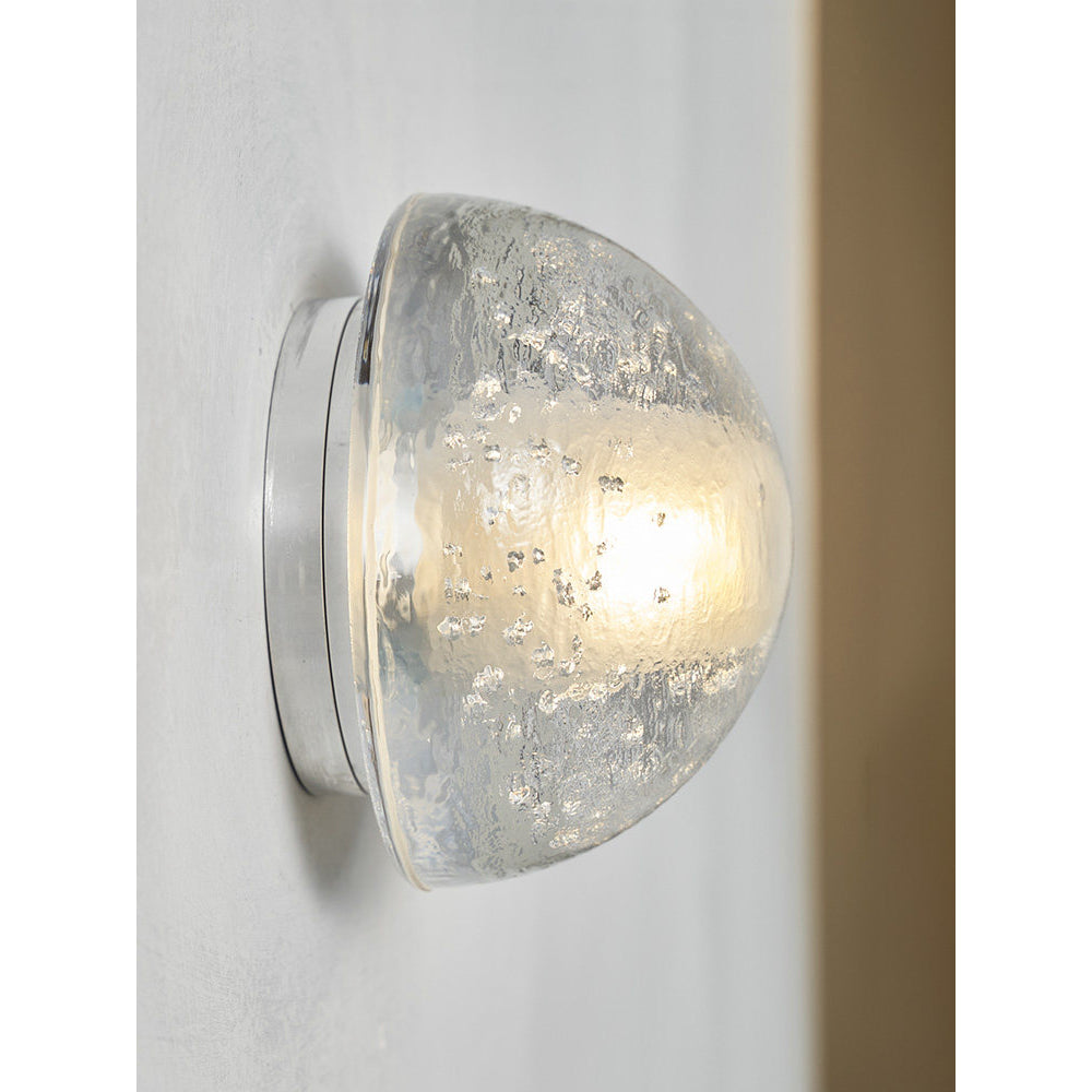 Angled view of a Clérachic wall lamp glowing warm beside a wooden panel, the clear dome sitting flush on its round plate.