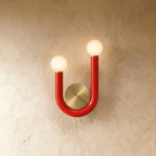 Clérachic U-shaped wall lamp in glossy red tube with two lit opal globes and a brushed brass backplate on a textured plaster wall.