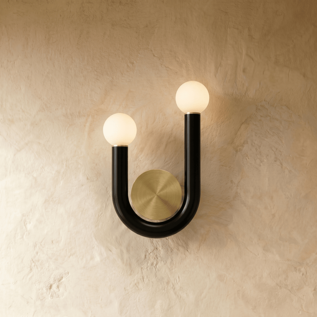 Clérachic U-shaped wall lamp in black tube with two lit opal globes and a brushed brass backplate, mounted on a limewash wall.