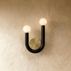 Clérachic U-shaped wall lamp in black tube with two lit opal globes and a brushed brass backplate, mounted on a limewash wall.