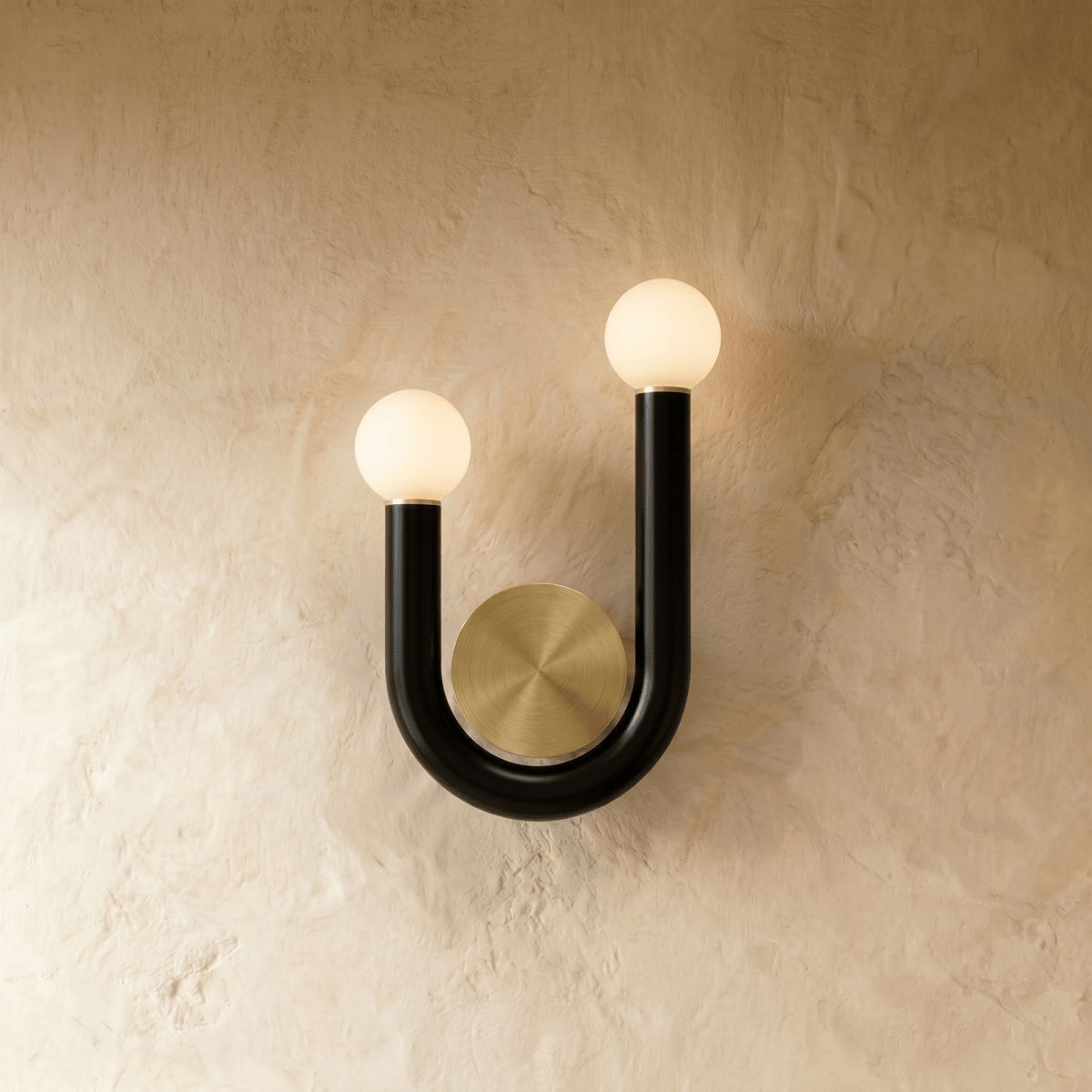 Clérachic U-shaped wall lamp in black tube with two lit opal globes and a brushed brass backplate, mounted on a limewash wall.