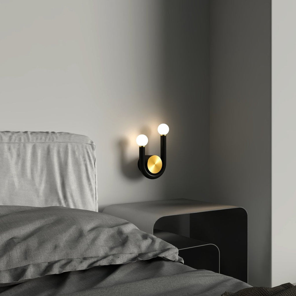Clérachic black U-shaped wall lamp with two glowing globes and a brass disc, fitted beside a grey bed above a low steel bedside table.