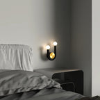 Clérachic black U-shaped wall lamp with two glowing globes and a brass disc, fitted beside a grey bed above a low steel bedside table.