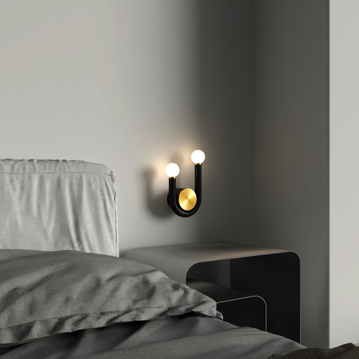 Clérachic black U-shaped wall lamp with two glowing globes and a brass disc, fitted beside a grey bed above a low steel bedside table.