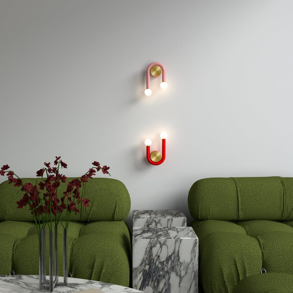 Clérachic pink and red U-shaped wall lamps, one inverted, hung on a pale wall above green boucle sofas and marble side tables.