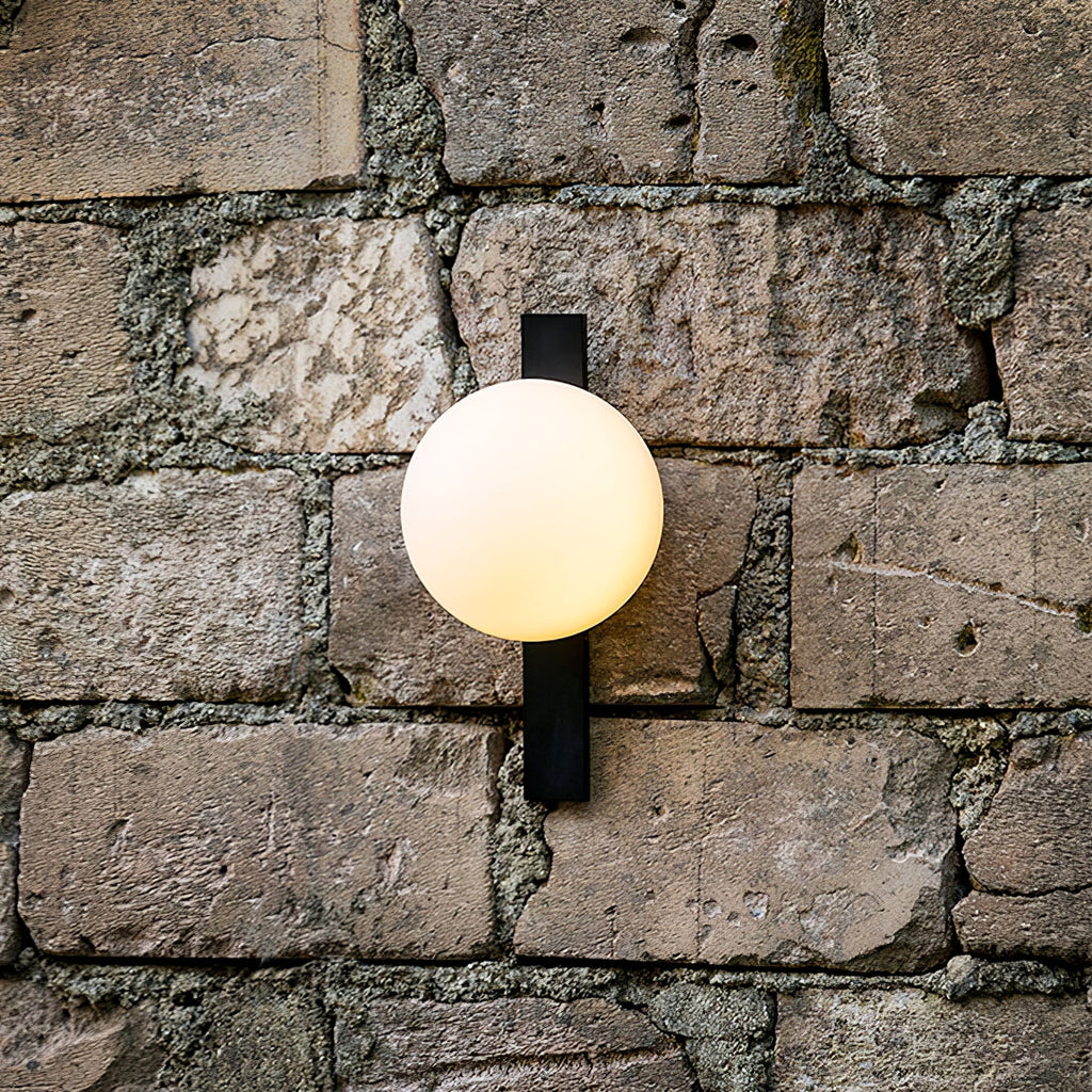 A lit cream globe on a black bar, this Clérachic wall light is mounted against a rough stone brick wall.