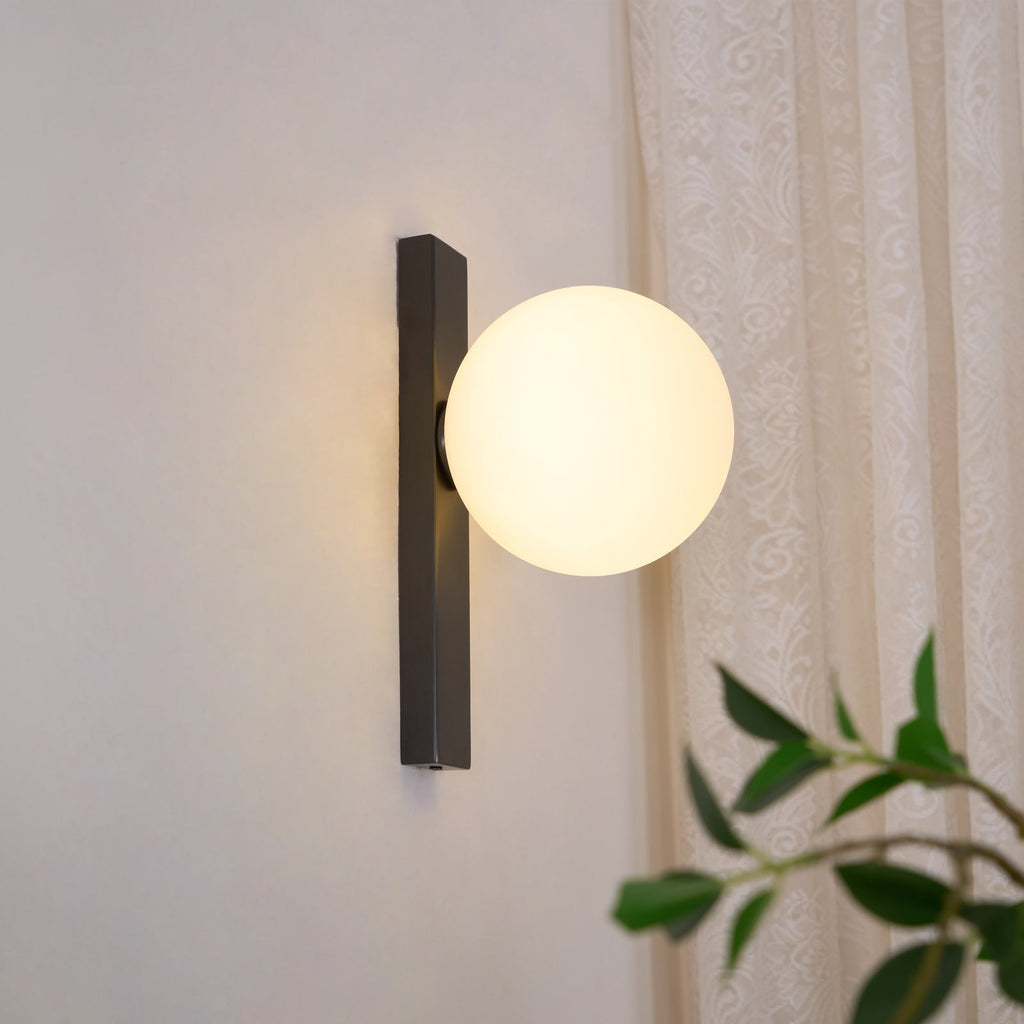 This Clérachic wall light lit on a pale wall, with a lace curtain behind and green leaves in the foreground.