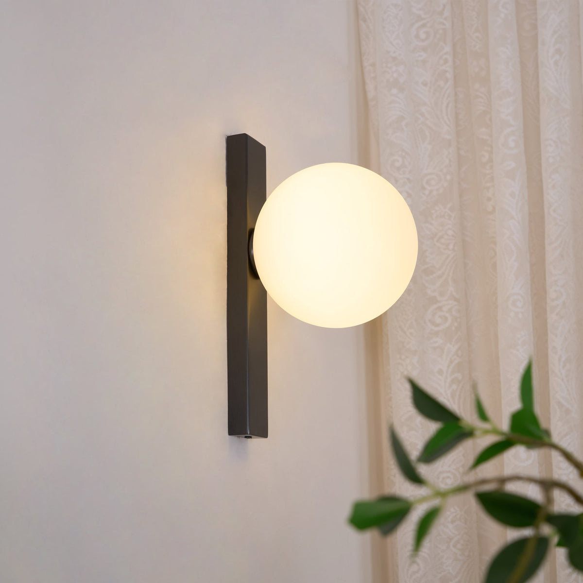 This Clérachic wall light lit on a pale wall, with a lace curtain behind and green leaves in the foreground.