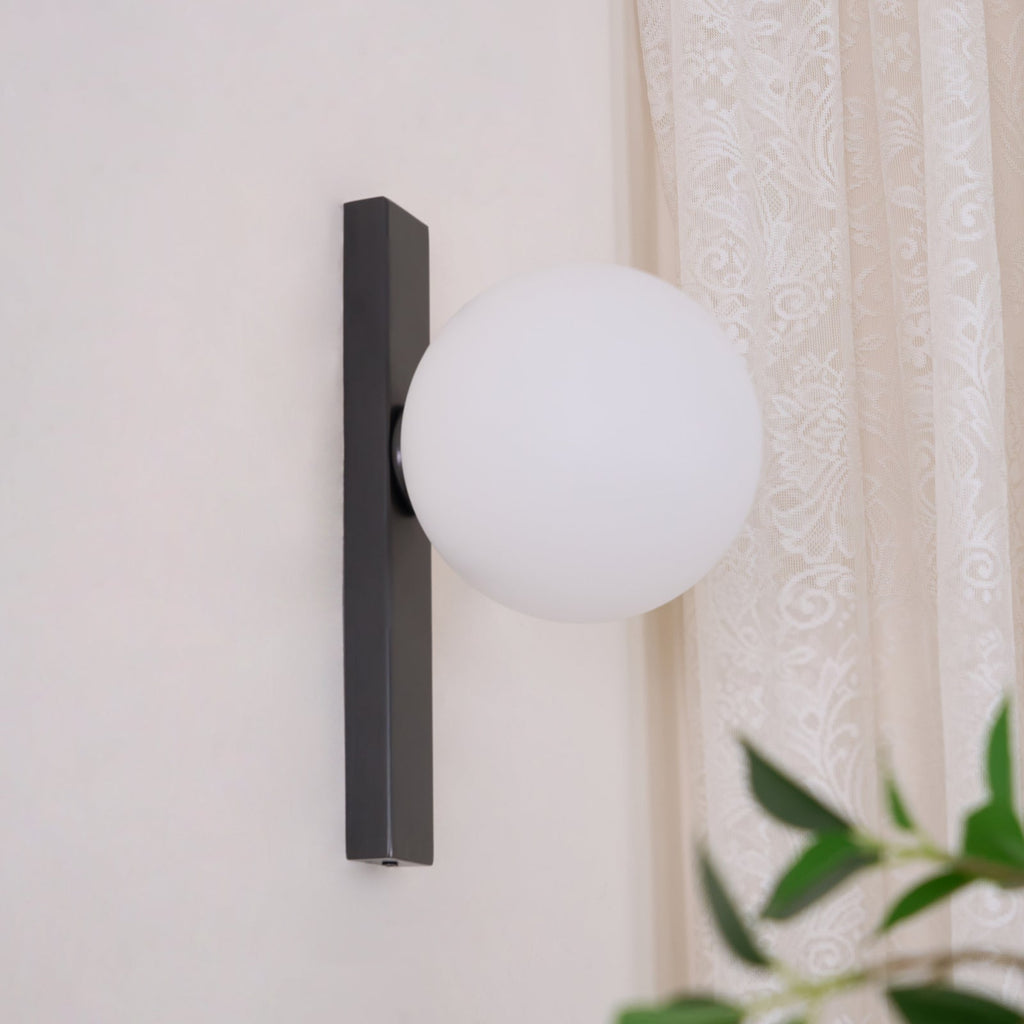 Unlit white globe on a black bar, this Clérachic wall light is mounted on a pale wall with a lace curtain beside it.