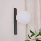 Unlit white globe on a black bar, this Clérachic wall light is mounted on a pale wall with a lace curtain beside it.