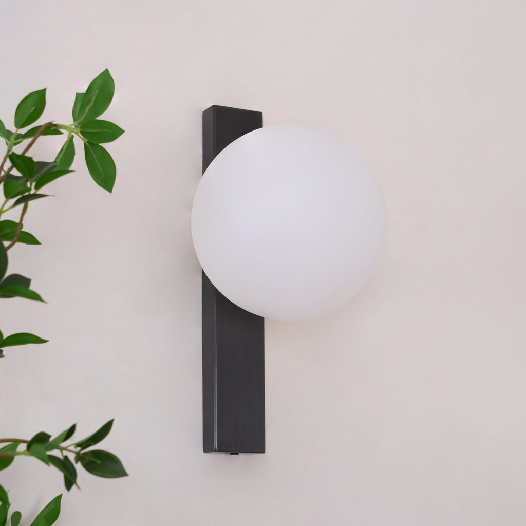 Clérachic wall light with a white globe on a vertical black bar, on a pale pink wall with leaves at the left.