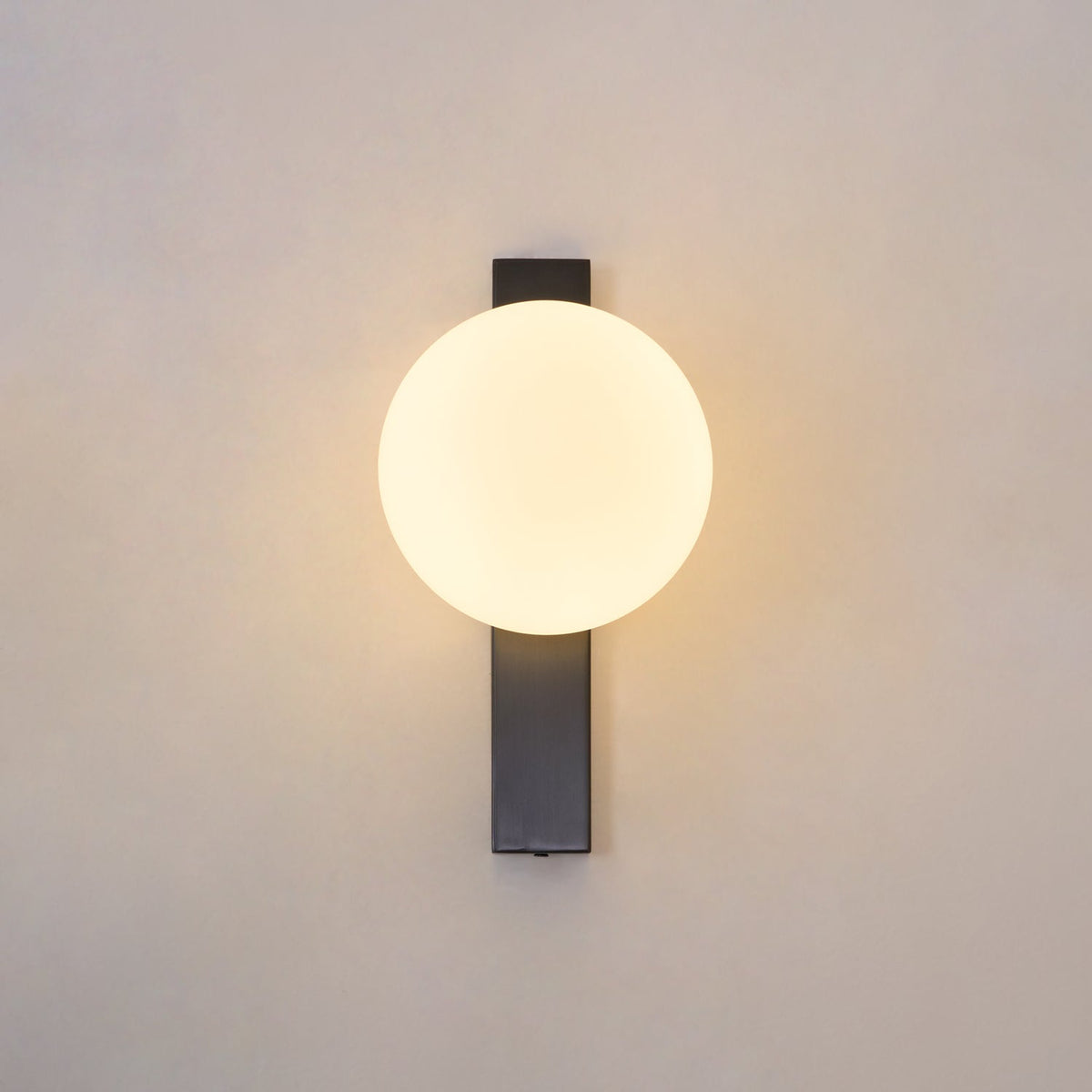 Centred on a warm beige wall, the glowing round globe of this Clérachic wall light sits on a slim black bar with a soft halo around it.