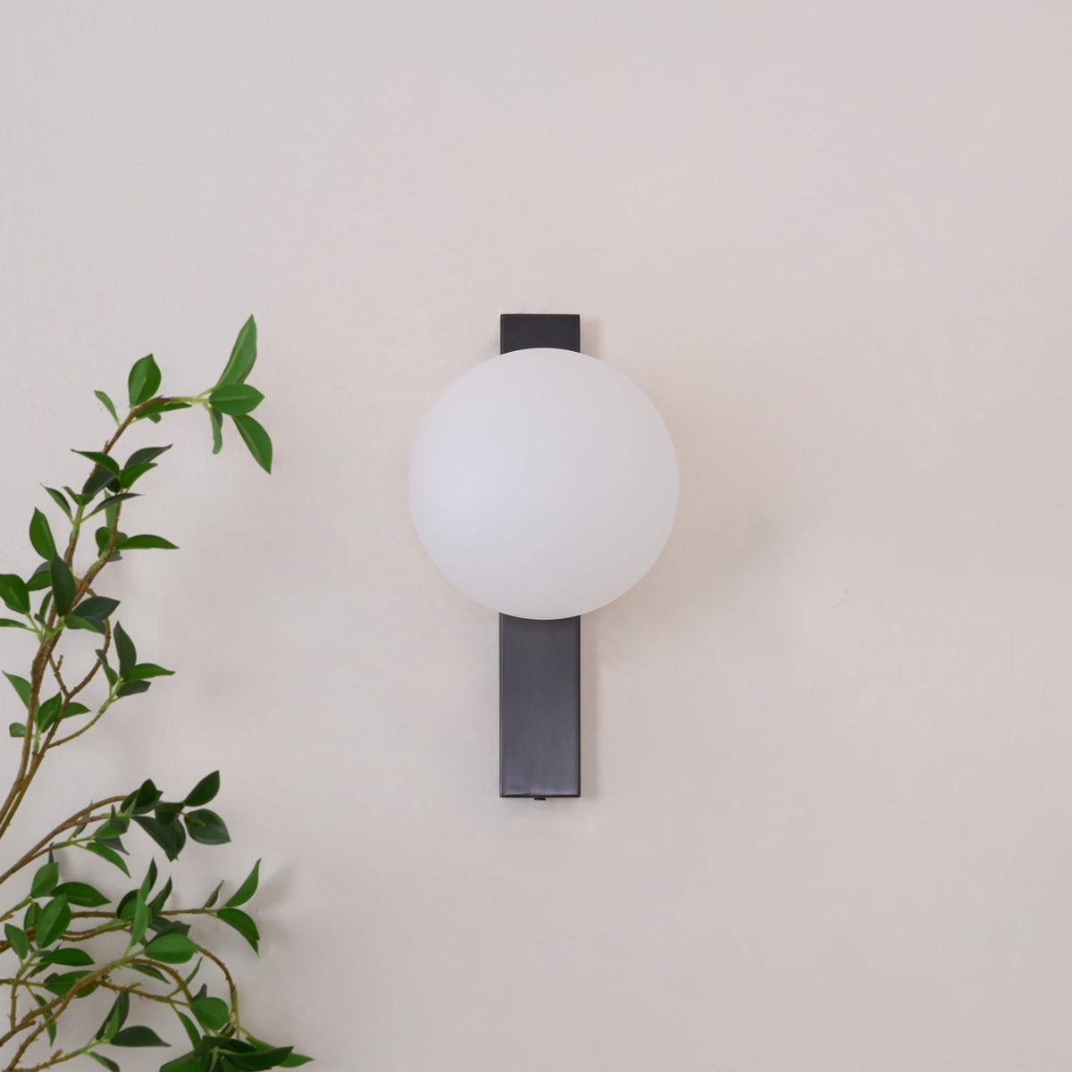 Small Clérachic wall light with an unlit white globe on a short black bar, beside a thin leafy branch on a cream wall.